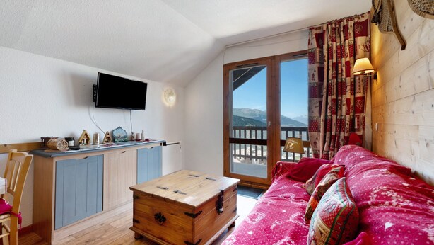 A cozy living room with a wooden coffee table, a red sofa, a television mounted on the wall, and a view of mountains through the window. - Apartment “Valmeinier Le Miras” with Mountain View, Private Terrace and Wi-Fi (Valmeinier)