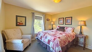A bedroom with a bed, a chair, a nightstand, a lamp, a window with curtains, and framed pictures on the wall. - Oceanfront Suite with Relaxing Tub & Private Bath - Rm 2 (Gabriola)