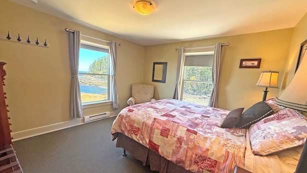 A bedroom with a large bed, two windows with curtains, a chair, a lamp, and a ceiling light. - Oceanfront Suite with Relaxing Tub & Private Bath - Rm 2 (Gabriola)