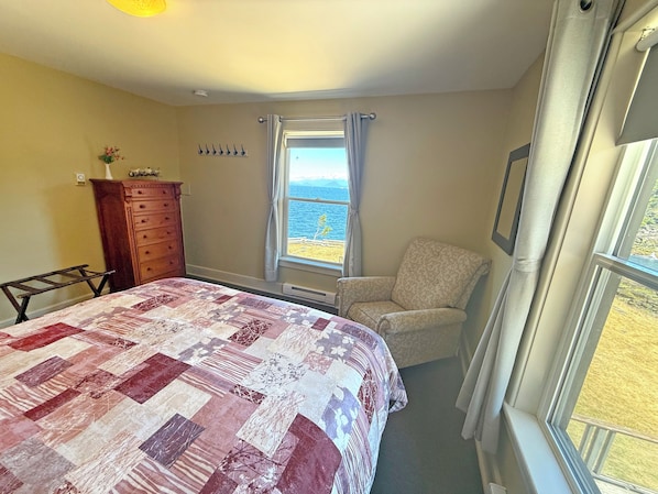 A bedroom with a bed, a chair, a dresser, and a window with a view of the sea.