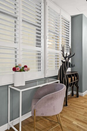 Ein Zimmer mit einem fensterartigen weißen Rahmen, einer grauen Wand, einem Holzboden, einem weißen Konsolentisch, einem violetten Sessel und einer Hirschskulptur.