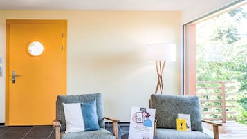 Ein Zimmer mit zwei grauen Sesseln, einem kleinen Tisch, einer gelben Tür und einem großen Fenster mit Blick auf Grünflächen.