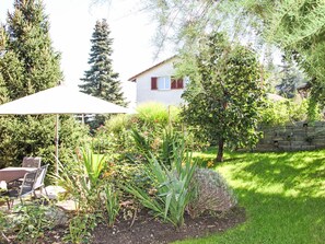 Ein Garten mit vielfältigen Pflanzen, eine Terrasse mit Tisch und Stühlen sowie ein Haus im Hintergrund.