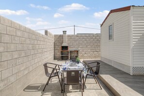 Um pátio com churrasqueira, mesa posta para refeições e um pequeno edifício branco.