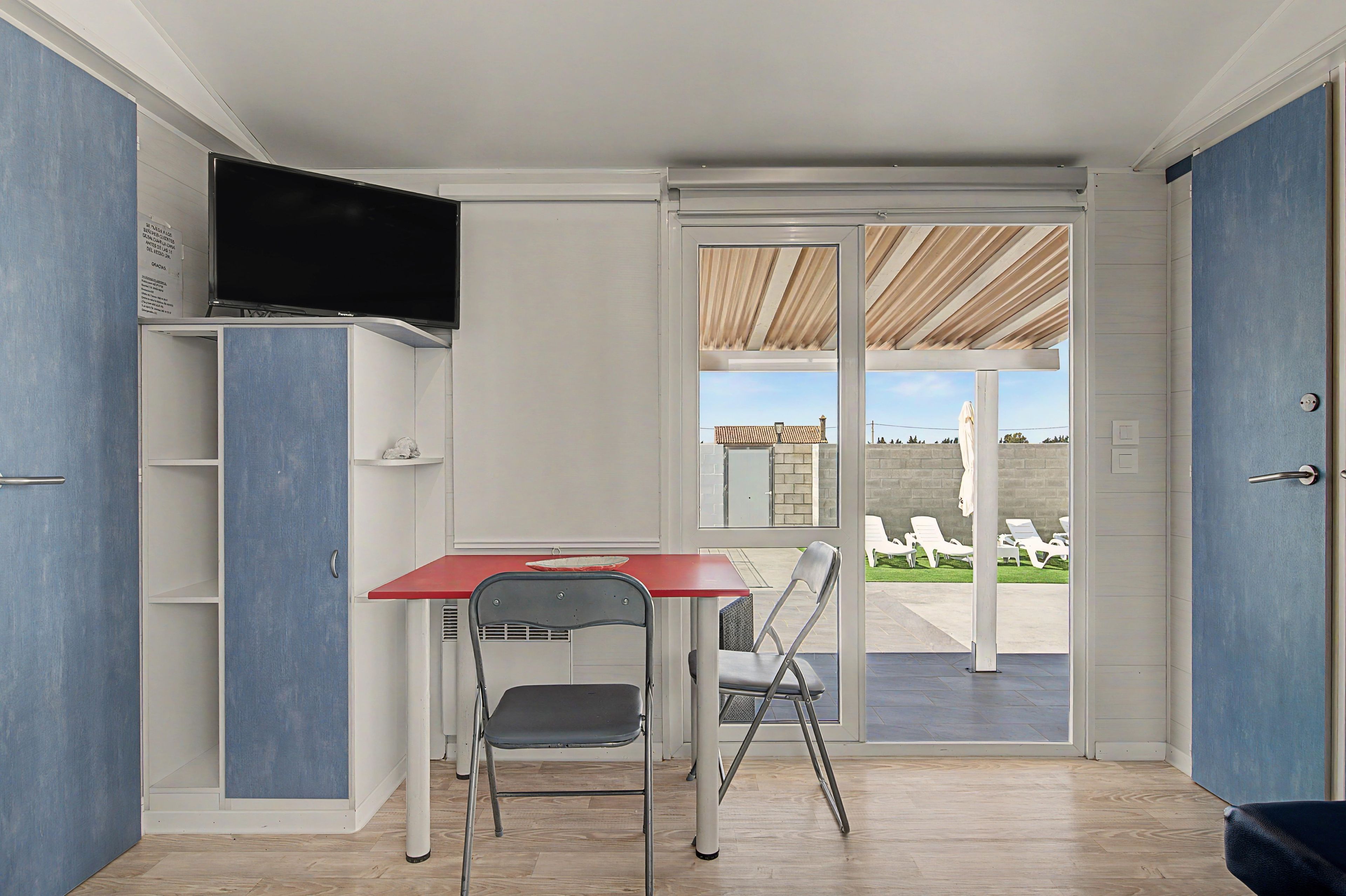 Une pièce compacte comprenant un coin cuisine, un espace repas avec une table et des chaises, et une porte-fenêtre coulissante donnant sur un espace extérieur aménagé avec des fauteuils de jardin.