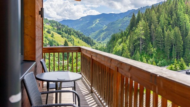 Un balcón de madera con una mesa y sillas, con vista a un paisaje montañoso cubierto de densos bosques.