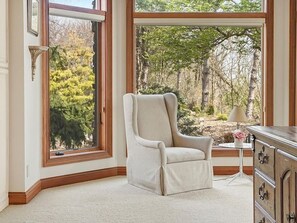 Una habitación con un ventanal grande, un sillón color beige, una mesita con una lámpara y un aparador de madera.