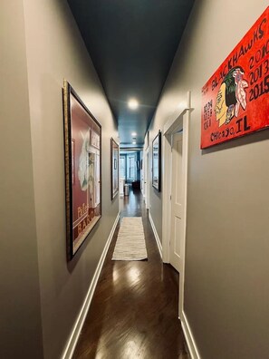 A hallway with framed artwork on the walls, a wooden floor, and a rug leading to a door.