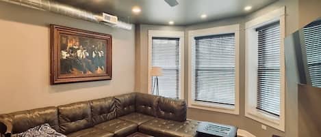 Ein Wohnzimmer mit einem ledernen Ecksofa, einem Glastisch, einem Deckenventilator und großen Fenstern mit Jalousien.