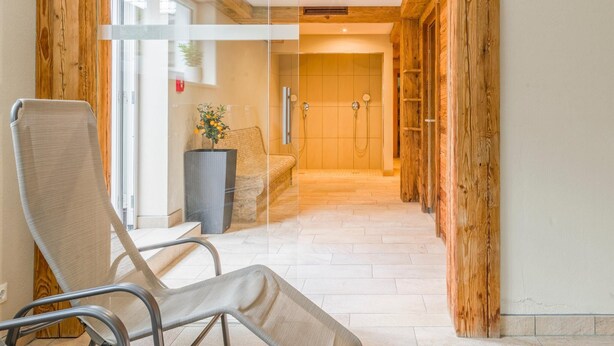 A modern bathroom with a glass shower enclosure, wooden beams, and a seating area with a chair and a small table. - Luxury apartment with wellness next to gondola (Brixen im Thale)