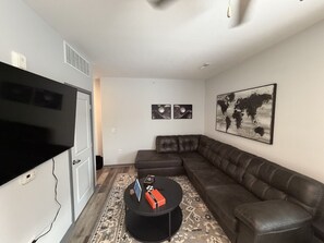 Ein Wohnzimmer mit einem großen Flachbildfernseher, einer dunkel ledernen Couch, einem runden Couchtisch mit einigen Gegenständen und einem Wandbild einer Weltkarte.