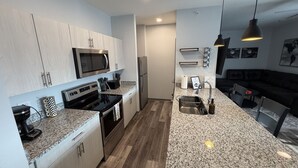 A modern kitchen with granite countertops, stainless steel appliances, and a dining area with a dark sofa. - Fast Lane Flat: Walk to Track, Dining & Parking (Speedway)