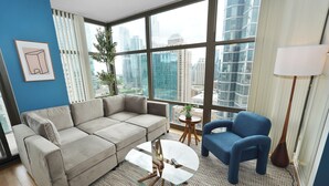 A modern living room with a sectional sofa, a round glass coffee table, a blue armchair, and a view of a cityscape through large windows.