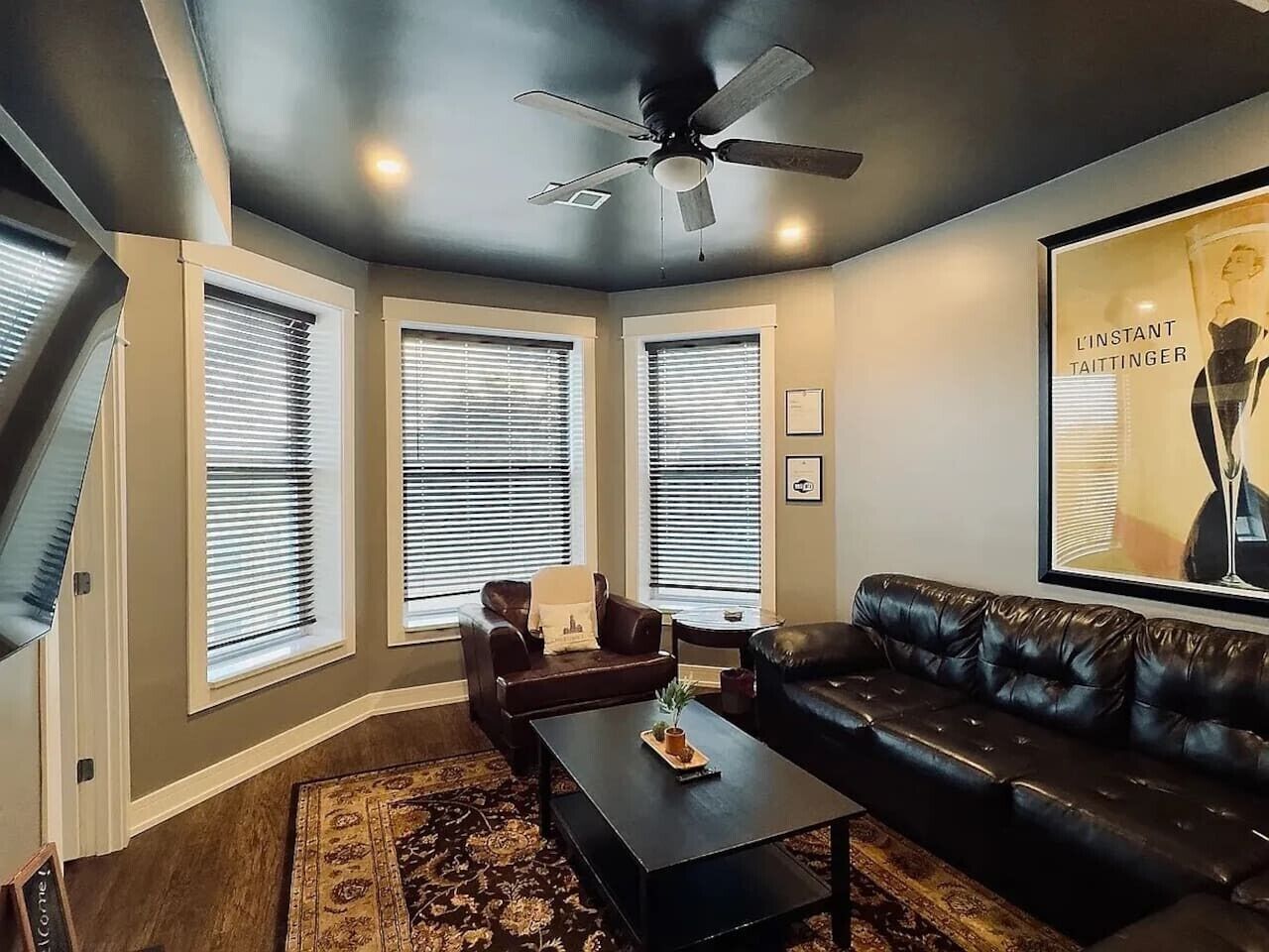 A living room with a black leather couch, a coffee table, and a ceiling fan. There are three windows with blinds, a rug, and a framed picture on the wall.