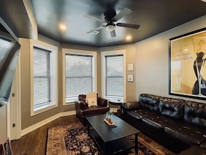 Ein Wohnzimmer mit einer schwarzen Ledersofa, einem Couchtisch und einem Deckenventilator. Es gibt drei Fenster mit Jalousien, einen Teppich und ein gerahmtes Bild an der Wand.