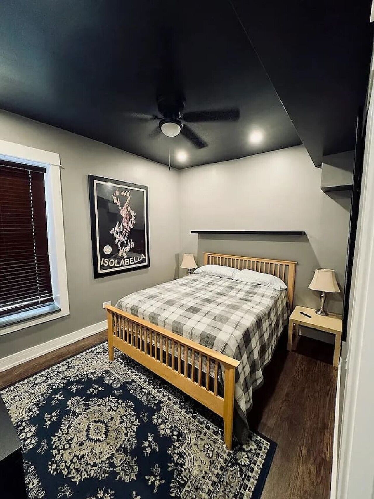 A bedroom with a bed, two bedside tables, a ceiling fan, a window with blinds, and a poster on the wall.