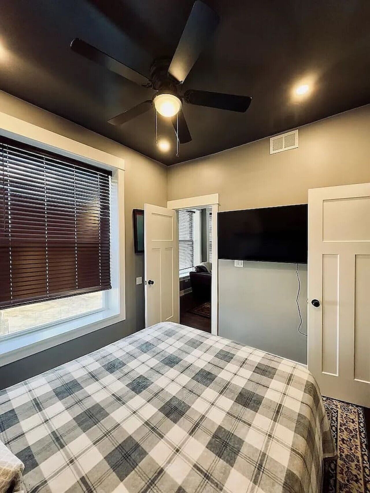 Une chambre dotée d’un ventilateur de plafond, d’un lit recouvert d’une couverture à carreaux, d’une fenêtre avec des stores et d’une porte donnant sur une autre pièce.