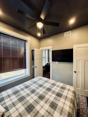 Ein Schlafzimmer mit Deckenventilator, einem Bett mit kariertem Überwurf, einem Fenster mit Jalousien und einer Tür zu einem anderen Raum.