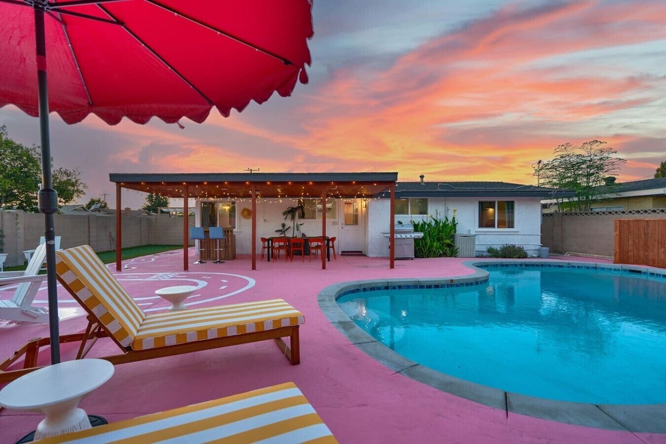 A backyard with a swimming pool, lounge chairs, a covered patio area, and a house in the background.