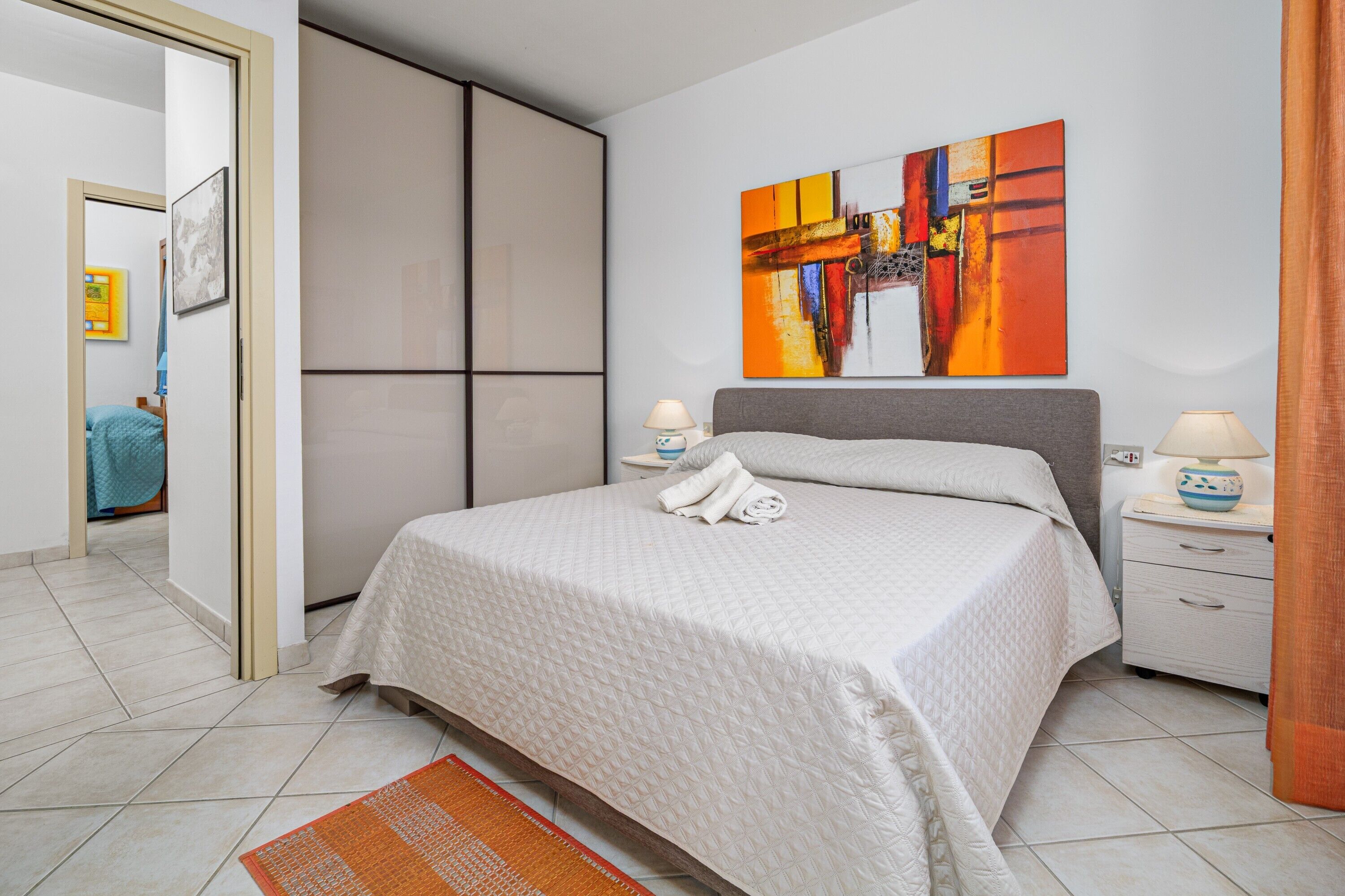 A bedroom with a bed, two bedside tables, a nightstand, a large abstract painting, and a wardrobe.