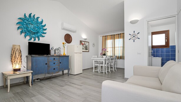 Una sala de estar con televisor, un aparador azul, sofá blanco, comedor con mesa y sillas blancas, y una cocina con azulejos azules.