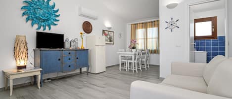Una sala de estar con televisor, un aparador azul, sofá blanco, comedor con mesa y sillas blancas, y una cocina con azulejos azules.