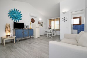 Una sala de estar con televisor, un aparador azul, sofá blanco, comedor con mesa y sillas blancas, y una cocina con azulejos azules.