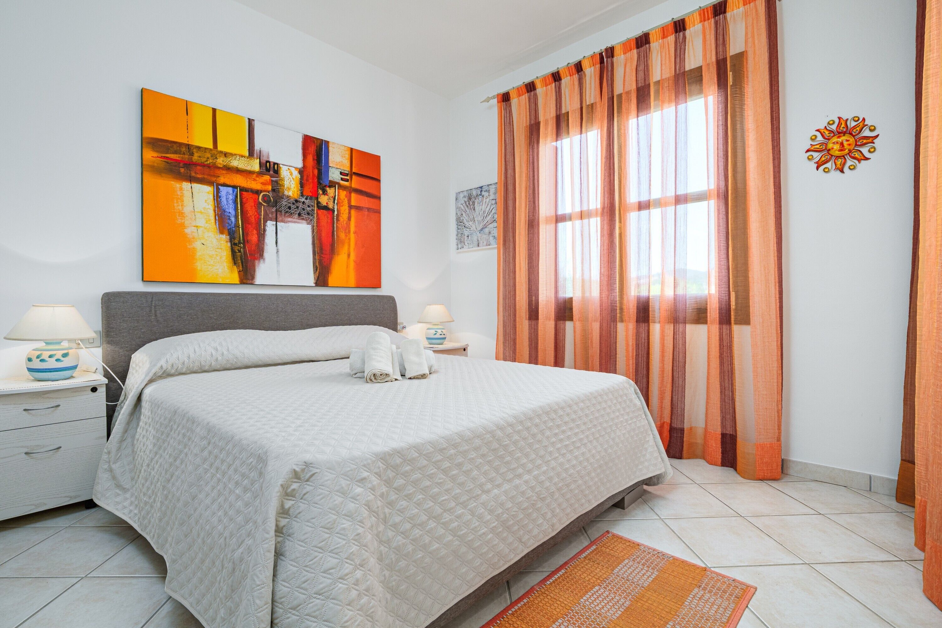 Ein ordentlich bezogenes Bett mit weißer Decke, ein Nachttisch mit Lampe und ein großes Fenster mit orangefarbenen Vorhängen.