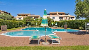 Una piscina con sillones de descanso y sombrillas, rodeada de setos y edificios.