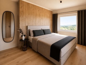 A modern bedroom with a large bed, a wooden headboard, a nightstand with a vase, and a view of a green landscape through a window. - Modern apartment for 16 people near the gondolas (Kirchberg in Tirol)