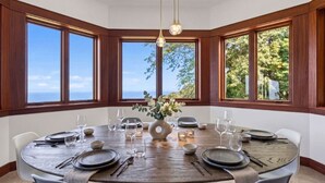 Ein runder Esstisch mit Stühlen, gedeckt für eine Mahlzeit, umgeben von großen Fenstern mit Meerblick.