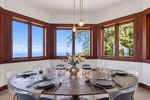 Ein runder Esstisch mit Stühlen, gedeckt für eine Mahlzeit, umgeben von großen Fenstern mit Meerblick.
