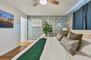 Une chambre d’hôtel avec un lit, un ventilateur de plafond, une plante, une fenêtre avec des rideaux et un téléviseur fixé au mur.