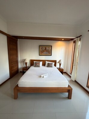 A spacious bedroom with a large bed, wooden headboard, two bedside tables with lamps, a painting on the wall, and a door on the left. - Room-a Quiet Room With Balcony in Nyuh K (Ubud)