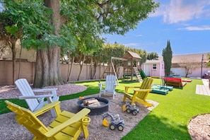 Ein Garten mit Adirondack-Stühlen, einer Feuerstelle, einem Spielhäuschen und einem kleinen Schuppen.