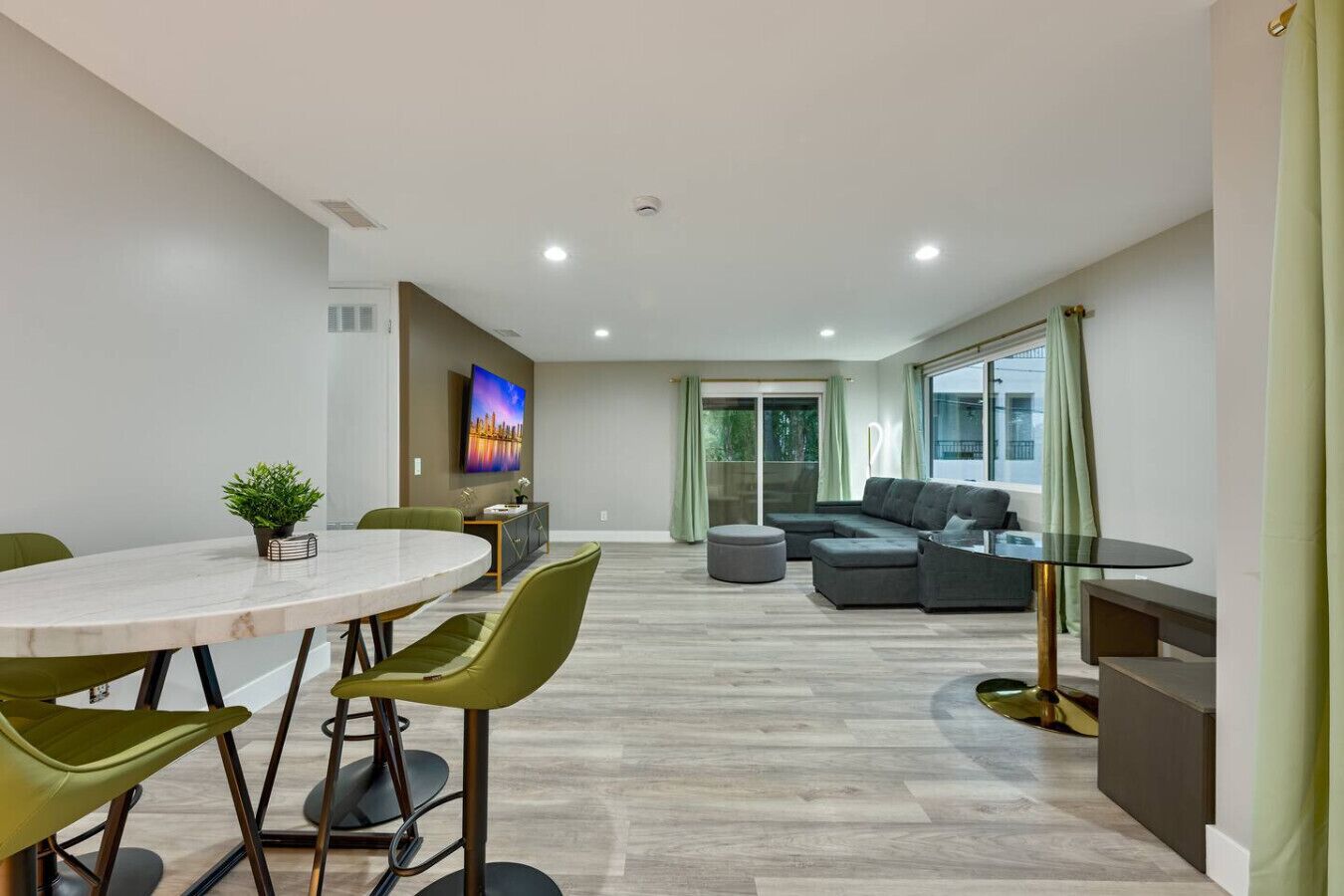 Un salon moderne comprenant un coin repas, un ensemble de canapés, un téléviseur fixé au mur et une porte-fenêtre coulissante donnant sur un balcon.