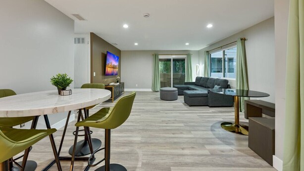 Una sala moderna con comedor, un conjunto de sofá, un televisor montado en la pared y una puerta corrediza de vidrio que da a un balcón.