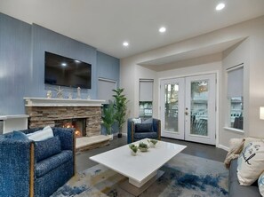 A living room with a stone fireplace, blue upholstered chairs, a white coffee table, and a flat-screen TV mounted on the wall.