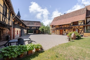 Une cour intérieure agrémentée de bâtiments traditionnels à colombages, d’une zone pavée et de plantes en pot.