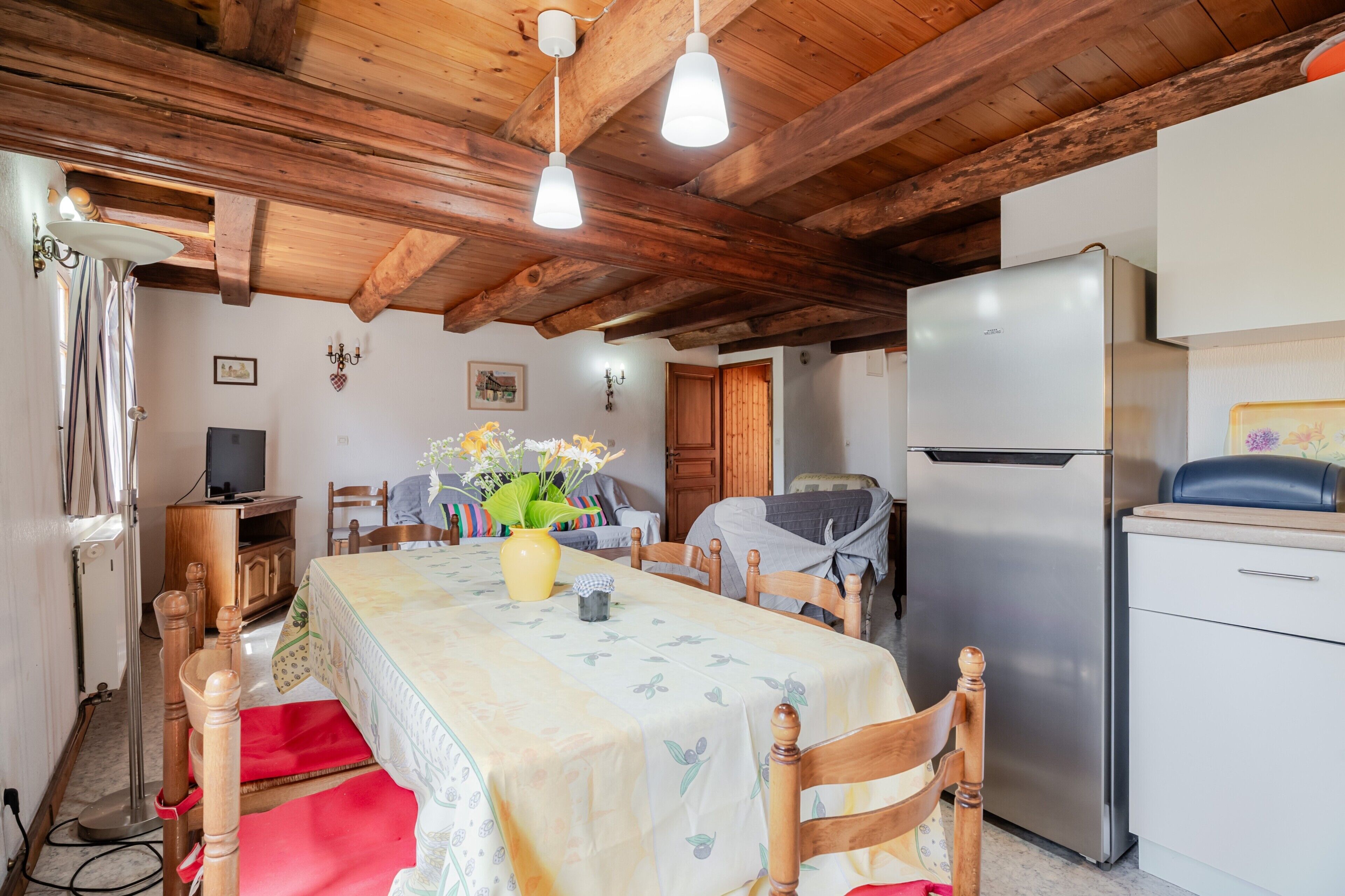 Un comedor con techo de madera, una mesa con mantel floral, sillas, un jarrón con flores, un refrigerador y un televisor sobre un mueble de madera.
