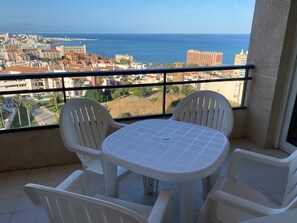 A balcony with a table and chairs, overlooking a coastal cityscape with buildings and the sea.