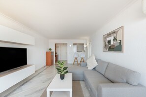 A modern living room with a flat-screen TV, a white coffee table, a grey sofa, and a minimalist kitchen in the background.