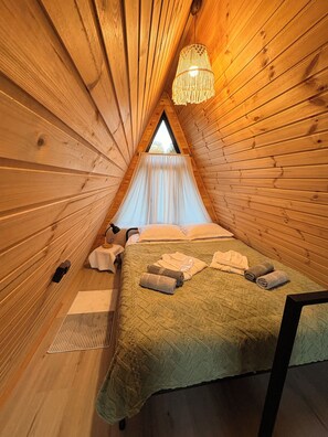 Une pièce triangulaire en bois, chaleureuse et accueillante, avec un lit, des serviettes, un luminaire suspendu et une fenêtre.