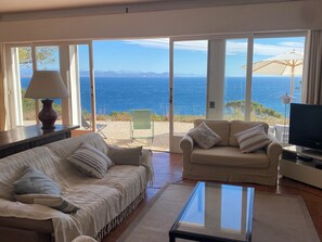 Ein Wohnzimmer mit einer Couch, einem Glastisch und einem Fernseher. Durch große Fenster ist der Meerblick zu sehen.