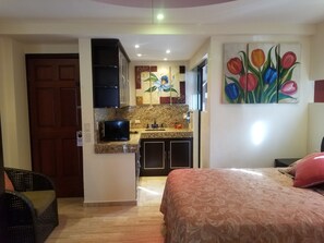 A compact hotel room with a bed, a small kitchenette, a television, and a painting of tulips on the wall. - PANORAMIC-2 SUITES Beachfront Condo Ocean Views 2-Full Bathrooms 2 Full Kitchens (Cancún)