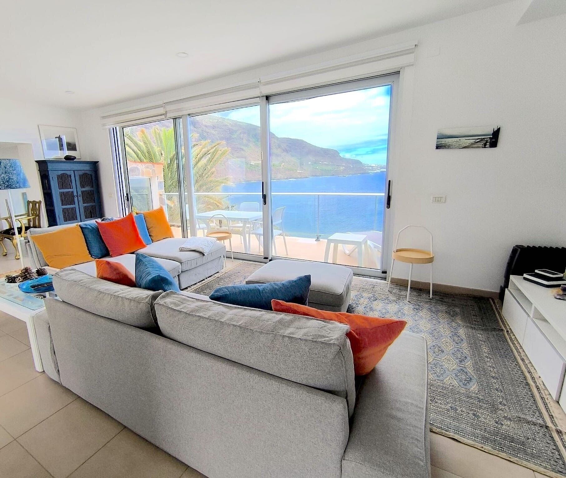 En moderne stue med en grå sofa, farverige puder, en glasdør der fører til en balkon med udsigt over hav og bakker.