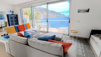En moderne stue med en grå sofa, farverige puder, en glasdør der fører til en balkon med udsigt over hav og bakker.
