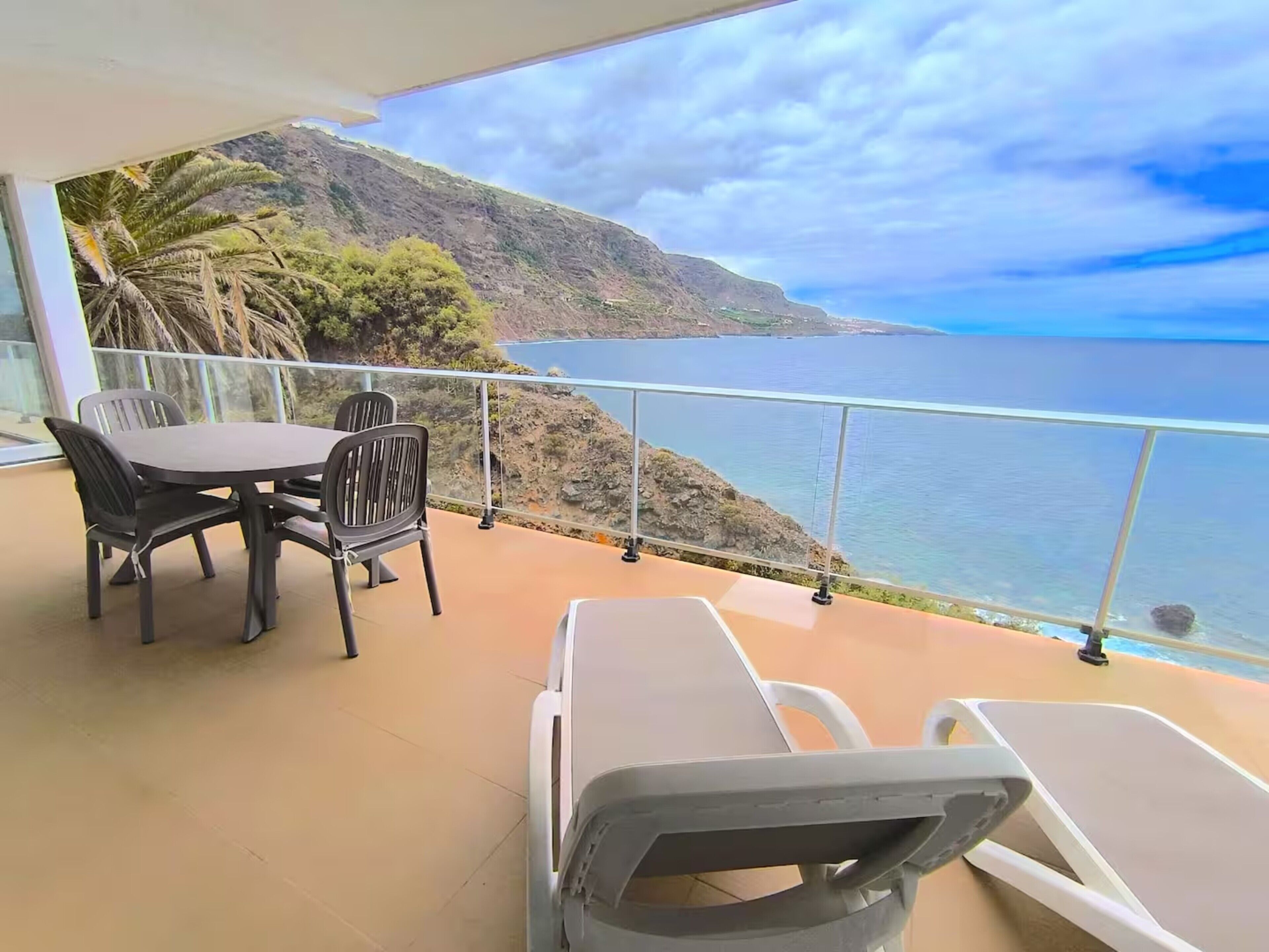 Ein Balkon mit Tisch und Stühlen, der auf eine Küstenlandschaft mit Blick auf das Meer und die Berge blickt.