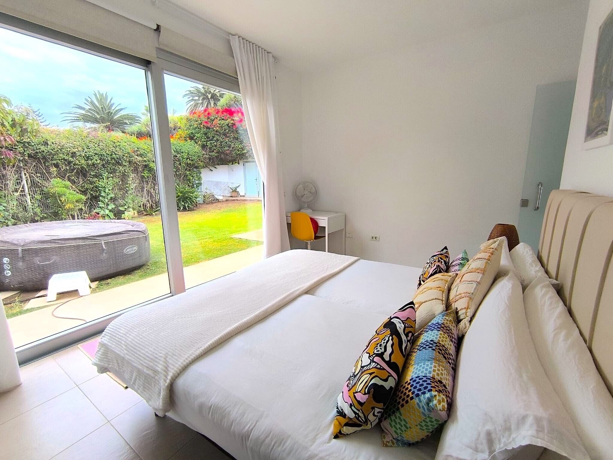 Un dormitorio con una cama grande, un escritorio pequeño, una silla y vistas a un jardín a través de un ventanal corredero.