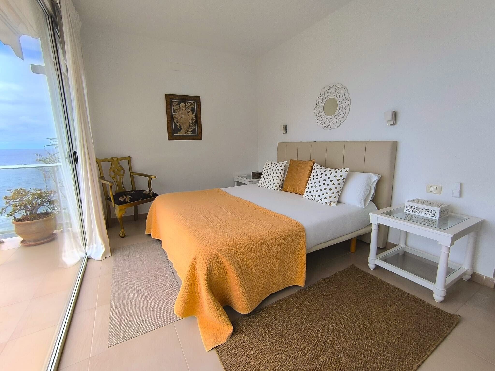 Een slaapkamer met een bed, nachtkastje, een potplant, een ingelijst kunstwerk en uitzicht op zee.